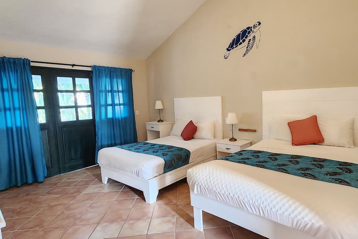 A room with two beds, a window with curtains, a wall-mounted artwork of a sea turtle, and a tiled floor.