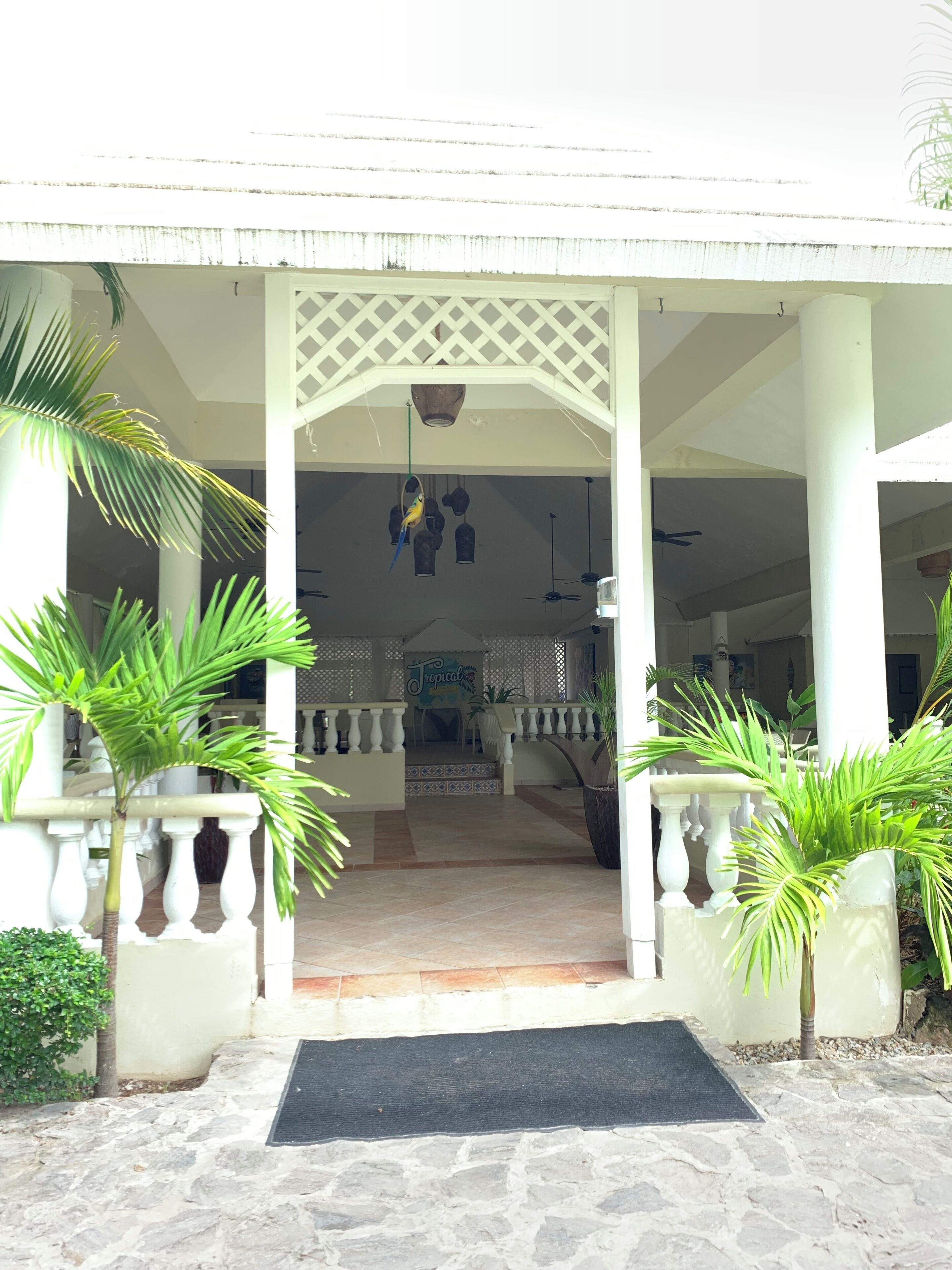 A covered entrance with a tiled floor, white columns, and a central doorway leading to a covered area with chairs and a sign reading "Tropical."