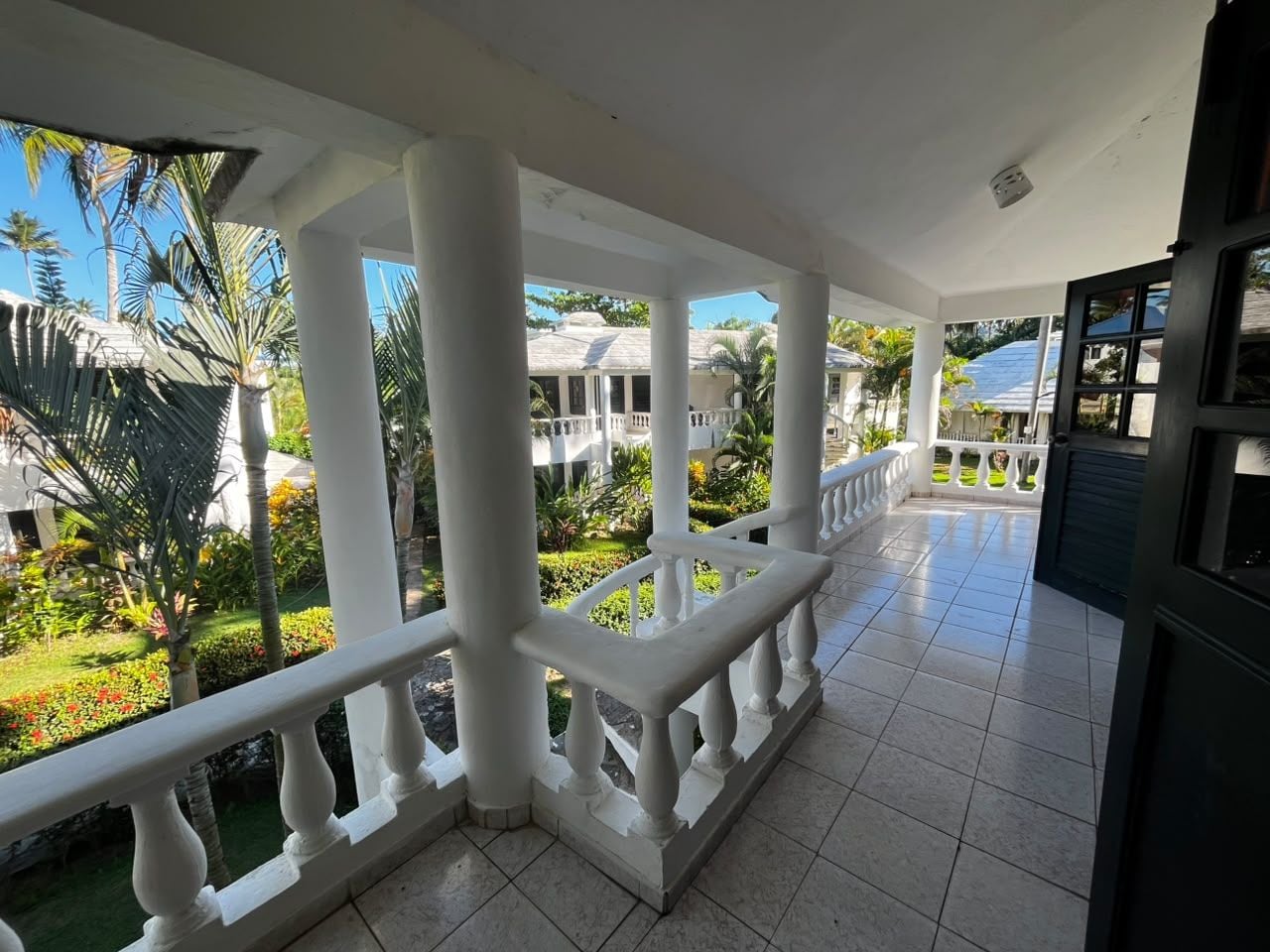 A covered balcony with white balustrades, a tiled floor, and a view of a garden with palm trees and a building in the background.
