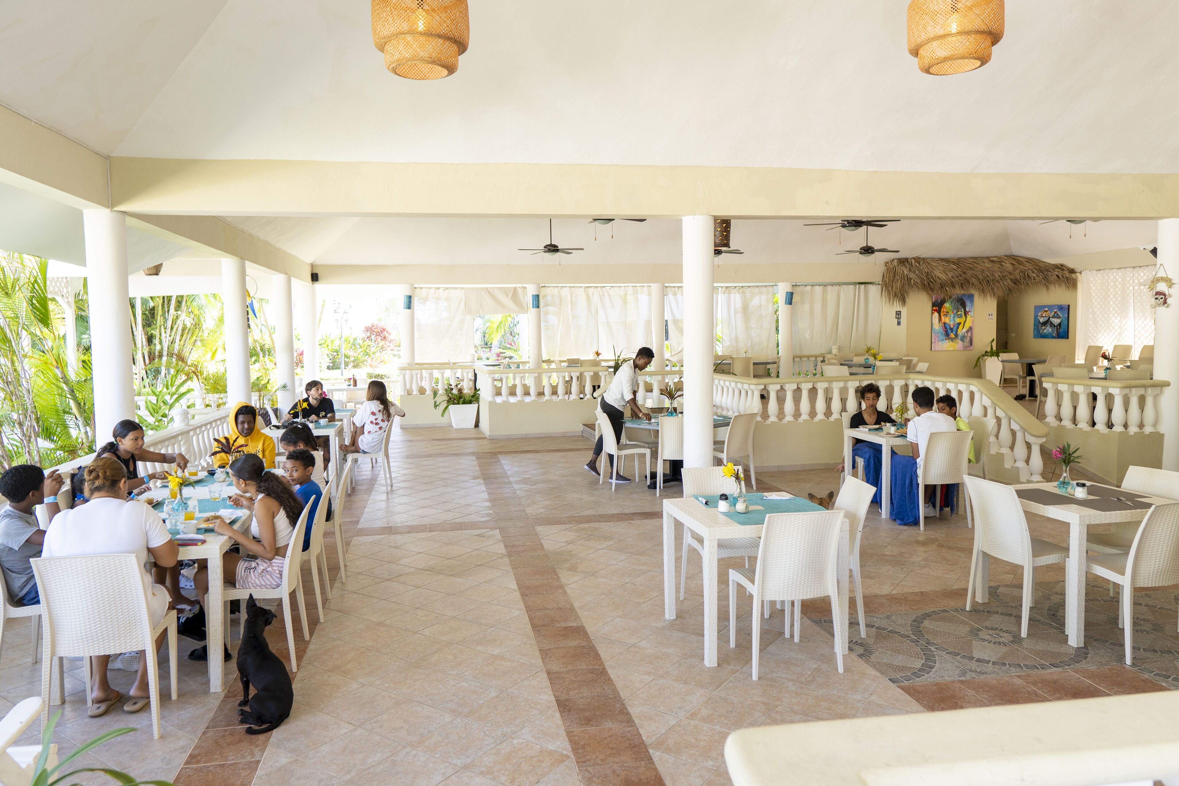 A spacious outdoor dining area with white furniture and thatched-roof tables, surrounded by potted plants and a view of greenery.