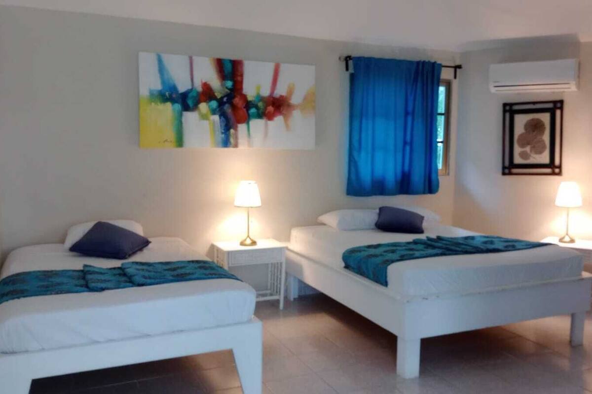 A room with two beds, each with a nightstand and a lamp. There is a colorful abstract painting on the wall, a blue curtain, and an air conditioning unit.