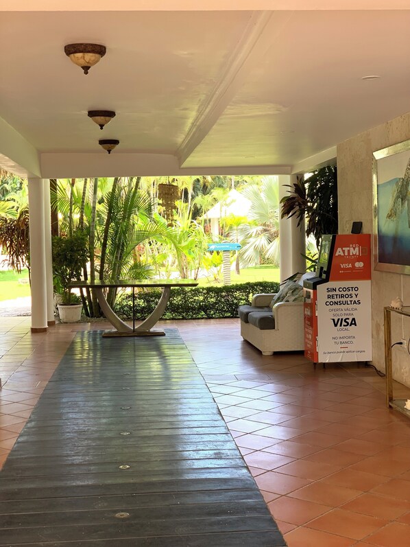 Un espacio al aire libre techado con suelo de baldosas, un banco y una mesa. Hay un cartel anunciando servicios Red ATM, Trinity y Visa.