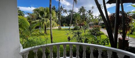 A balcony with a white balustrade overlooking a well-maintained garden with palm trees and a pool.
