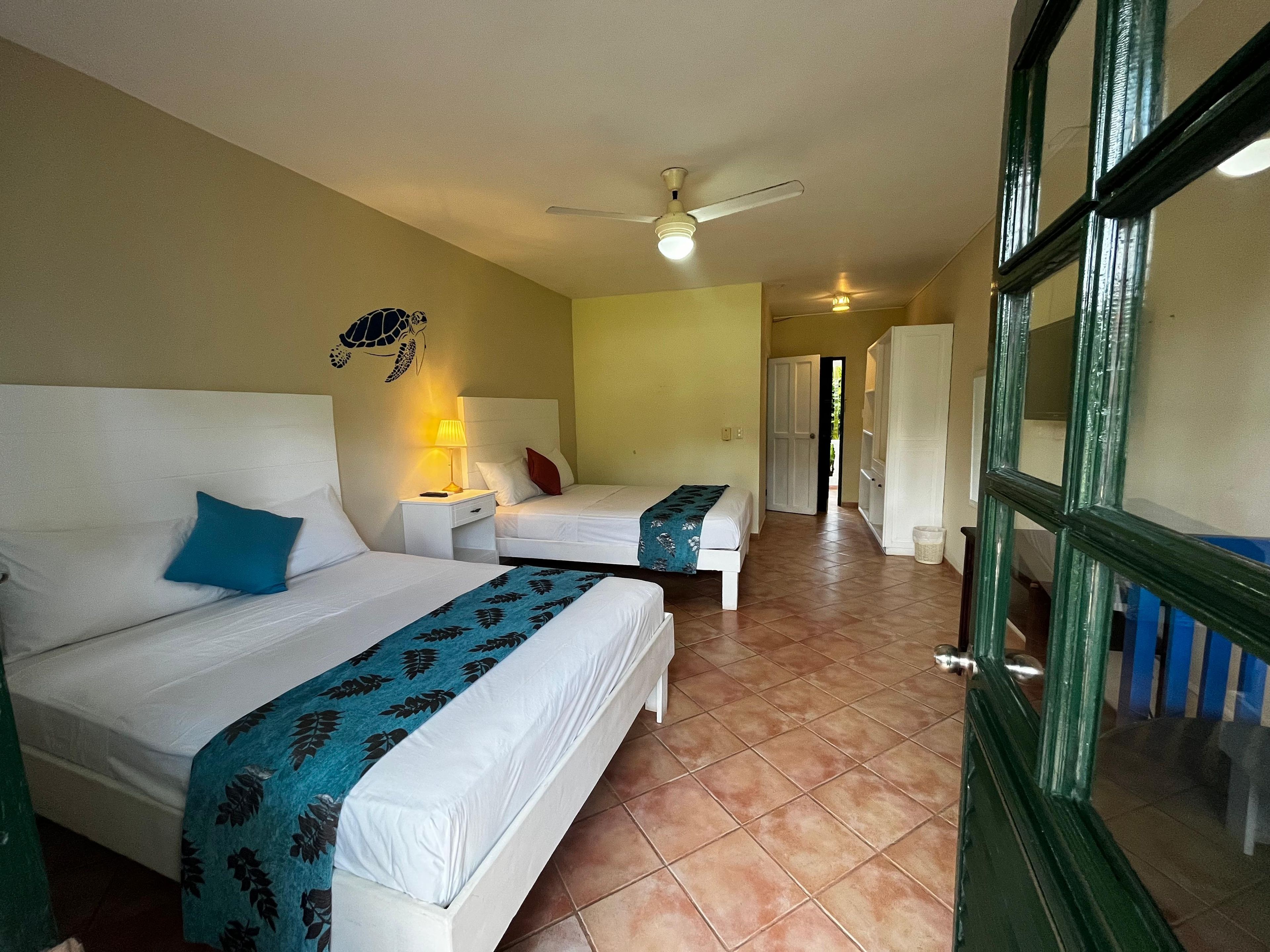 A hotel room with two beds, a ceiling fan, and a view of the corridor.