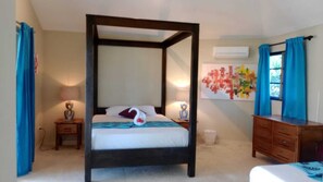 A four-poster bed with a swan towel, a wooden nightstand with a lamp, a dresser, and a wall with colorful artwork. - Hotel Bahia Punta Bonita (Las Terrenas)