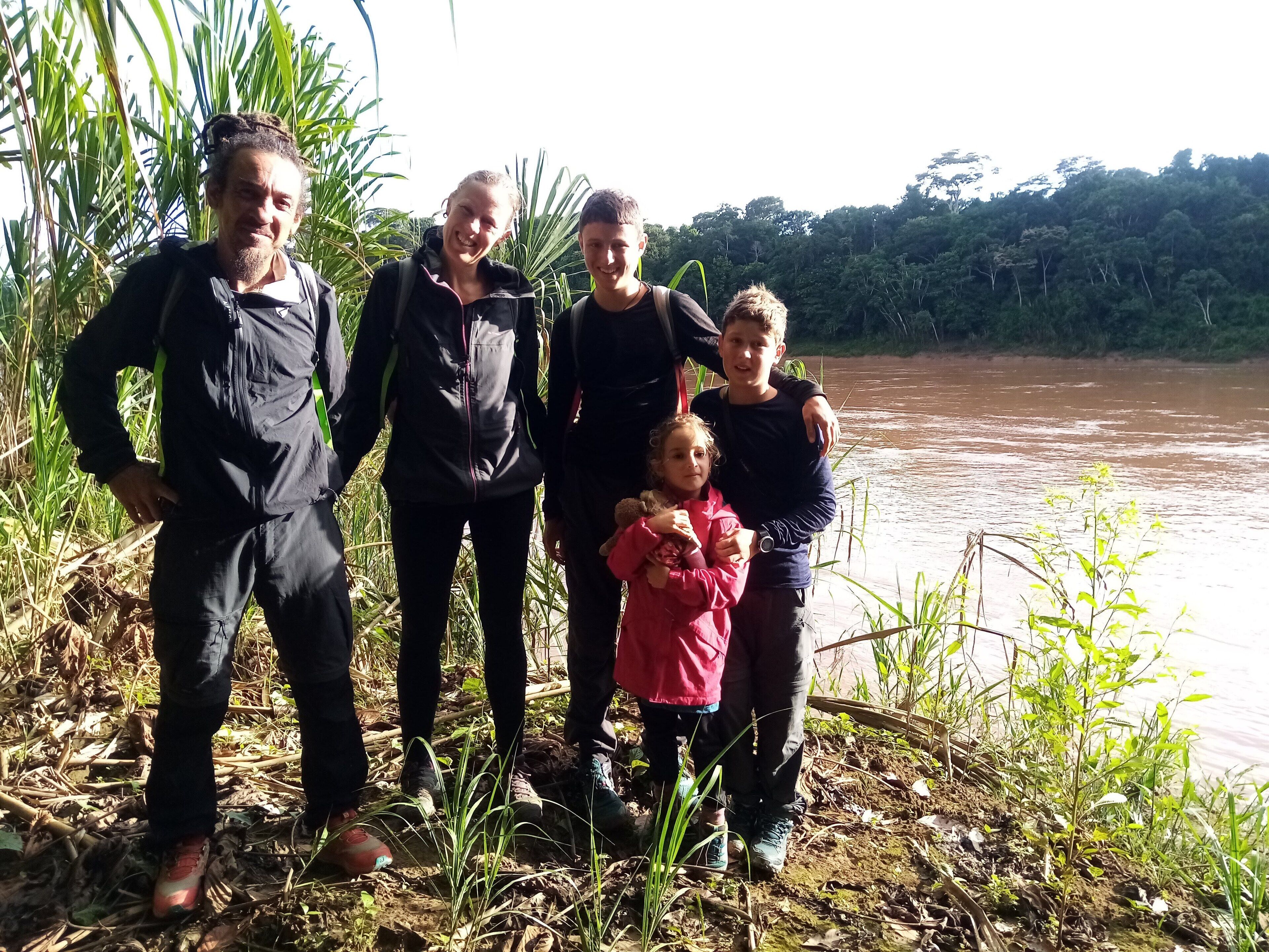 A group of people standing by a riverbank with vegetation and a forest in the background.