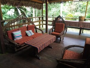 A wooden outdoor seating area with a thatched roof, a cushioned sofa, a striped table, and wicker chairs.