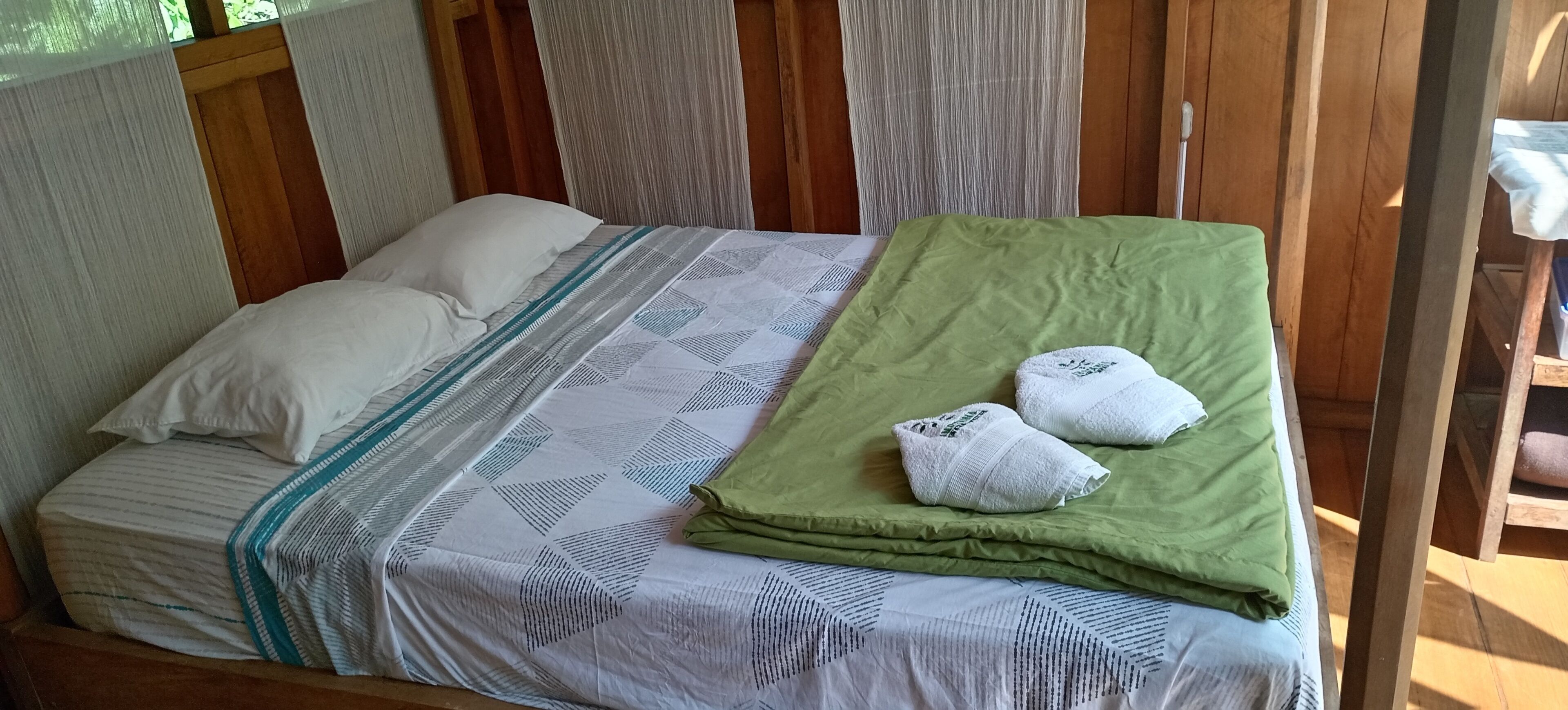 Una cama individual con una colcha estampada y almohadas, una manta verde y dos toallas sobre la cama.
