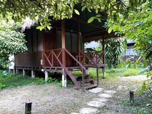 A wooden structure with a thatched roof, surrounded by lush greenery and a gravel path leading to it.