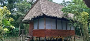 A wooden cabin with a thatched roof, surrounded by lush greenery.