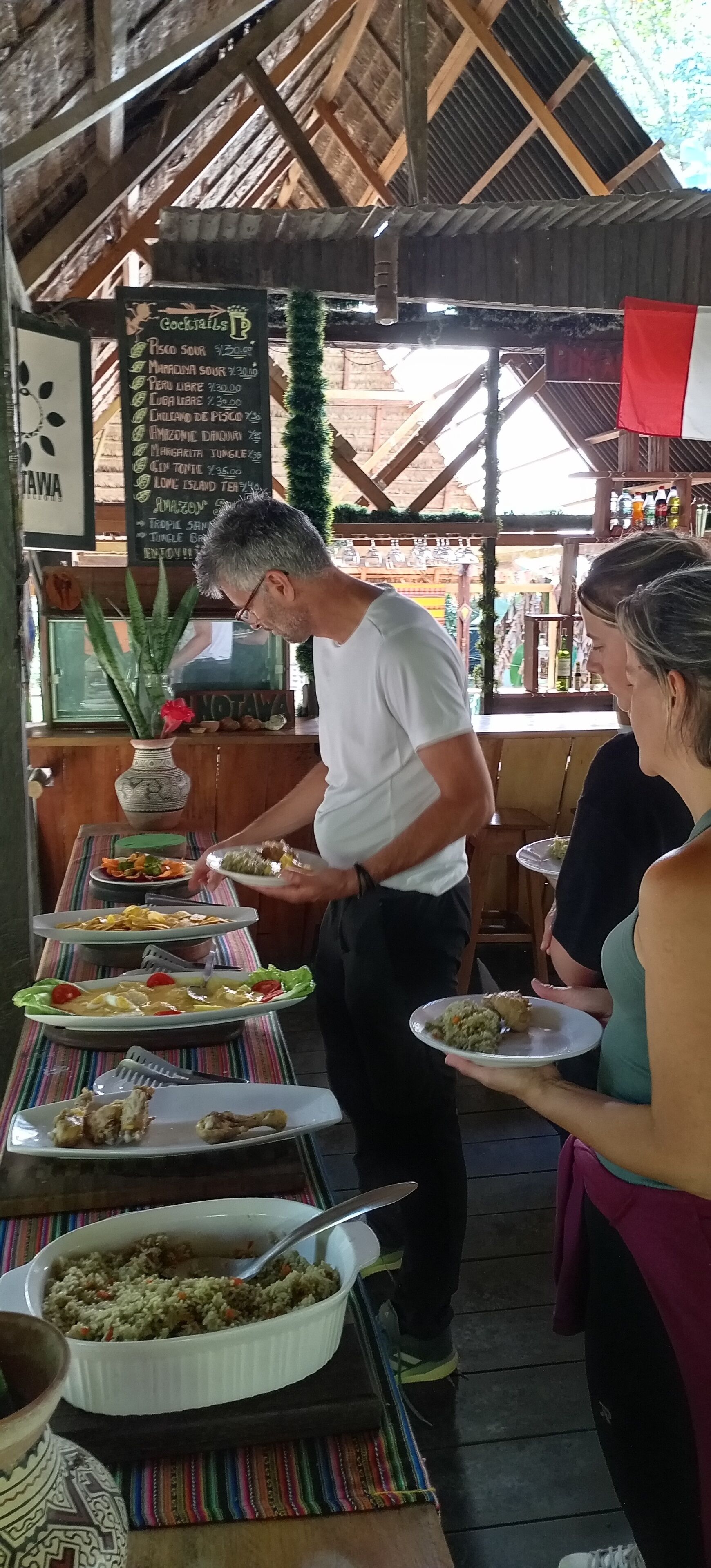 A rustic outdoor dining area with wooden beams and a corrugated metal roof. There are multiple dishes on a buffet table, including salads, meats, and a large bowl of rice. A signboard with cocktail options is visible in the background.