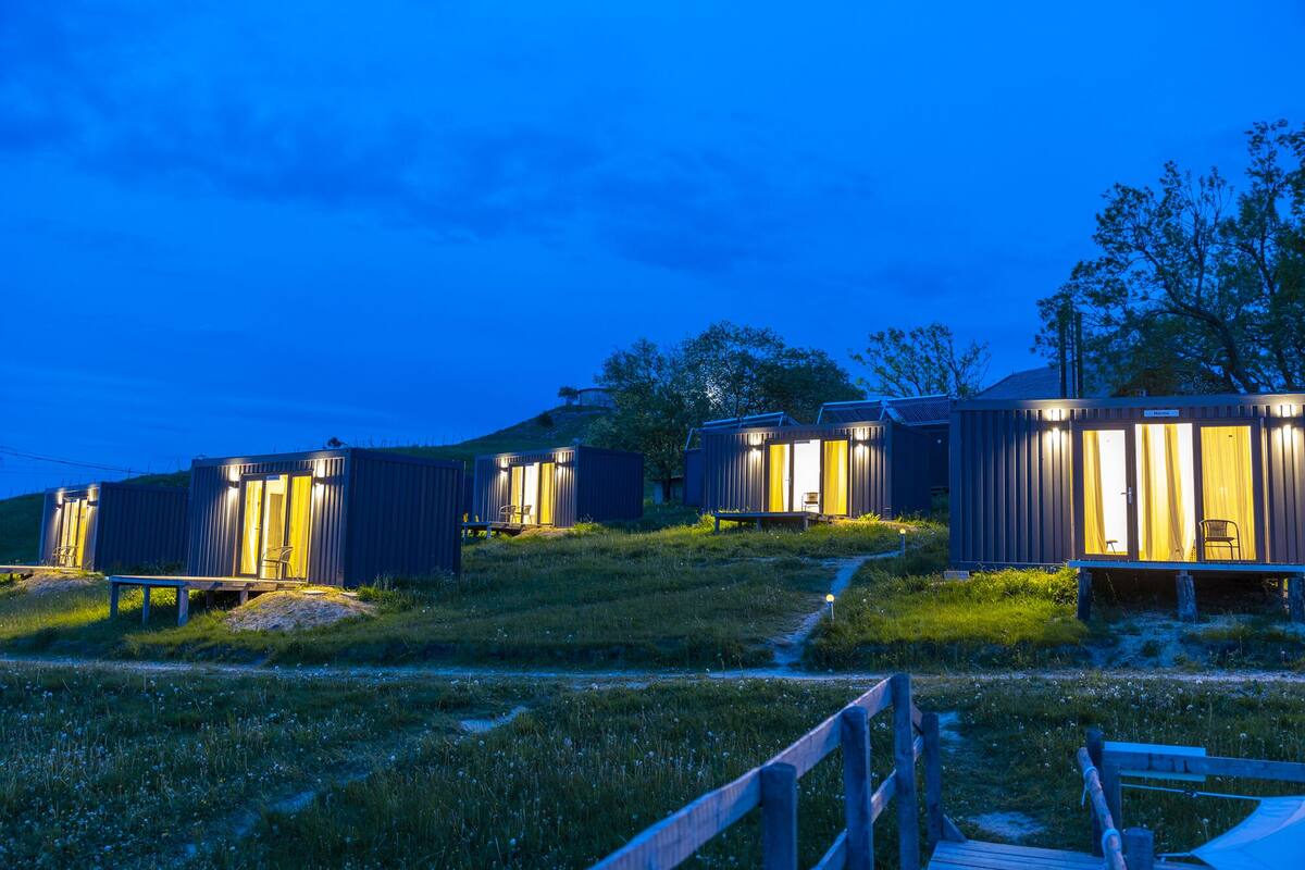 Een rij moderne, compacte accommodaties met openslaande ramen, elk voorzien van een klein terrasje en een stoel, gelegen voor een achtergrond van bomen en een heldere lucht.