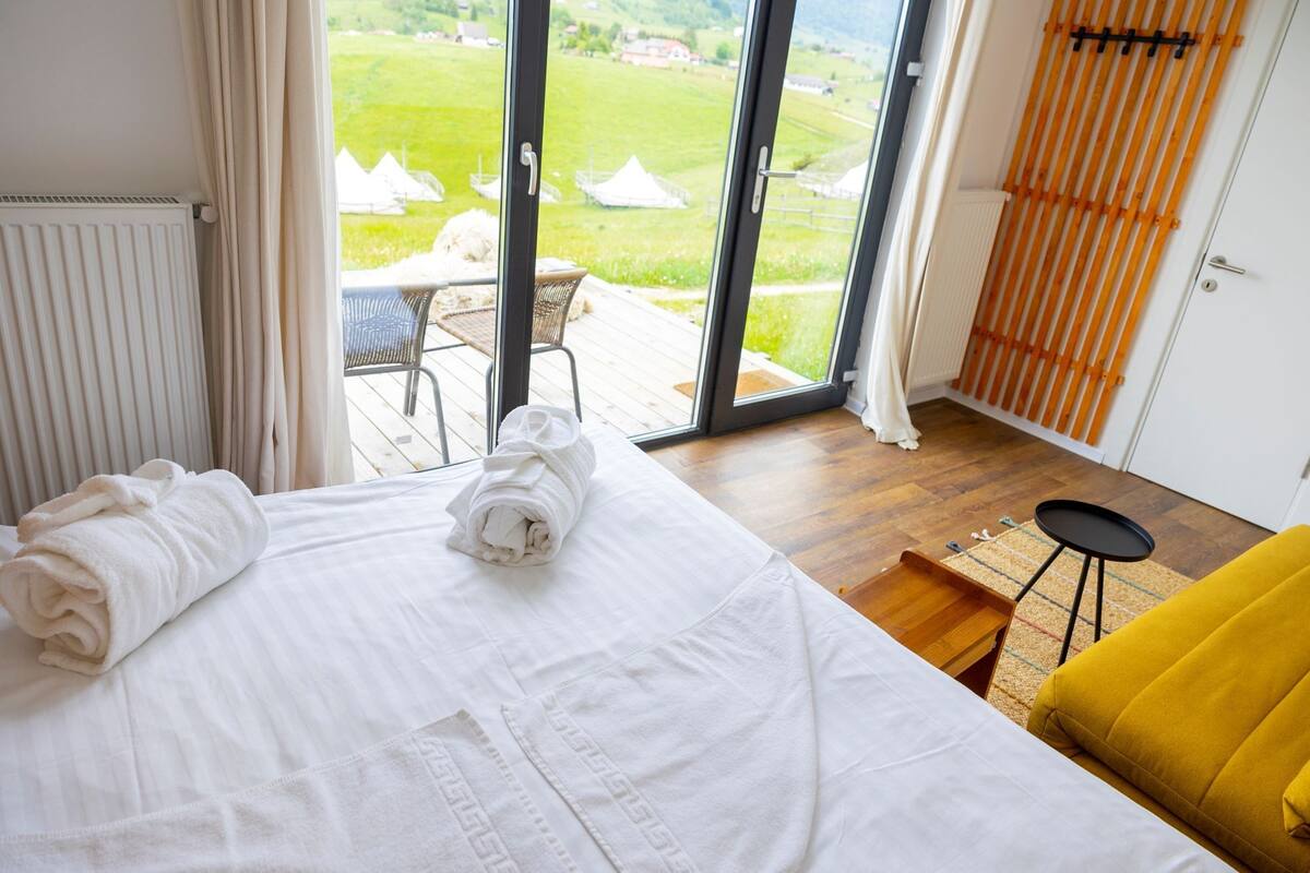 Een hotelkamer met een bed, een gele stoel, een kleine tafel en uitzicht op een groen landschap door grote glazen deuren.