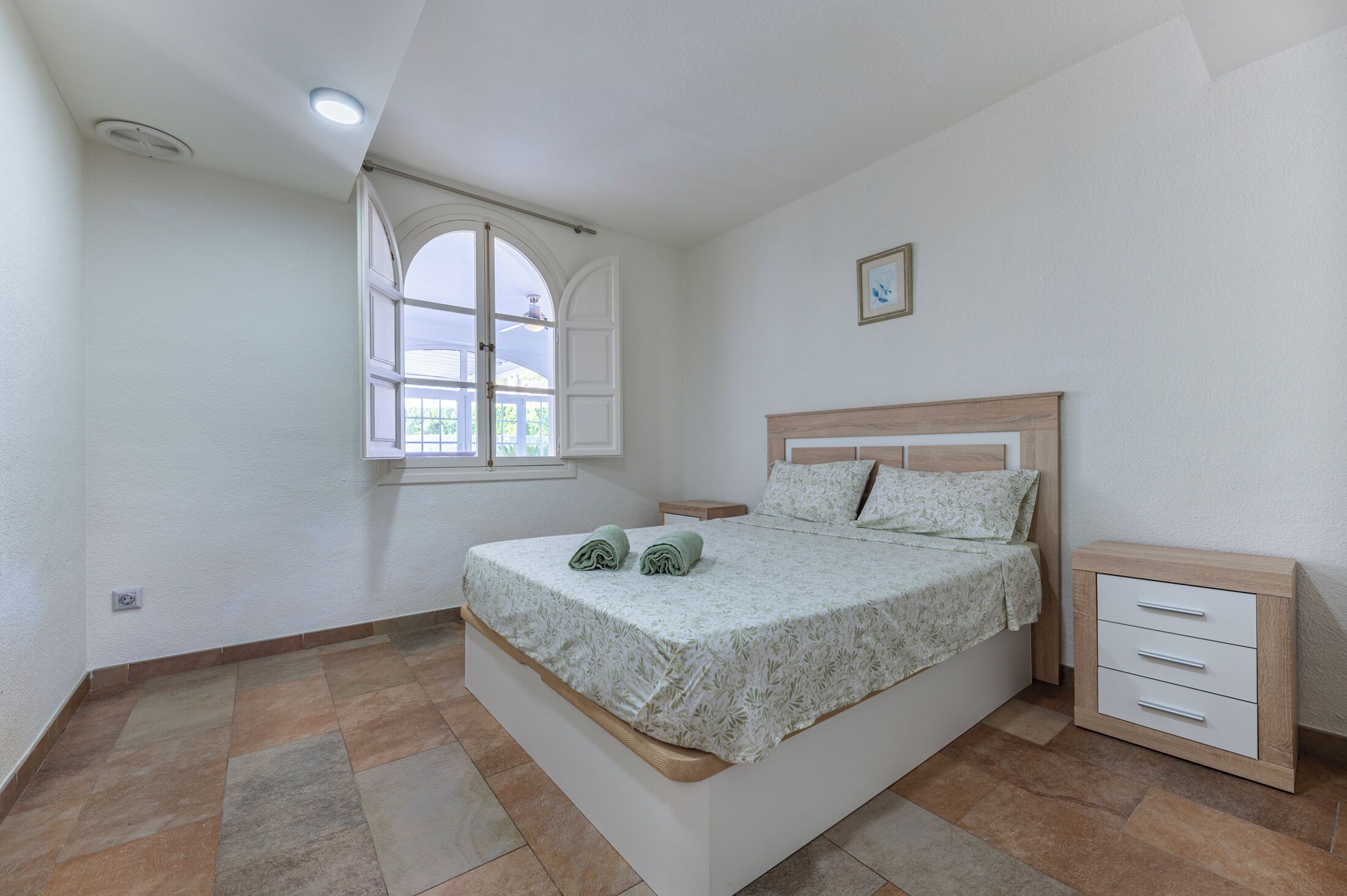 A bedroom with a bed, a nightstand, a drawer unit, a window with curtains, and a ceiling light.