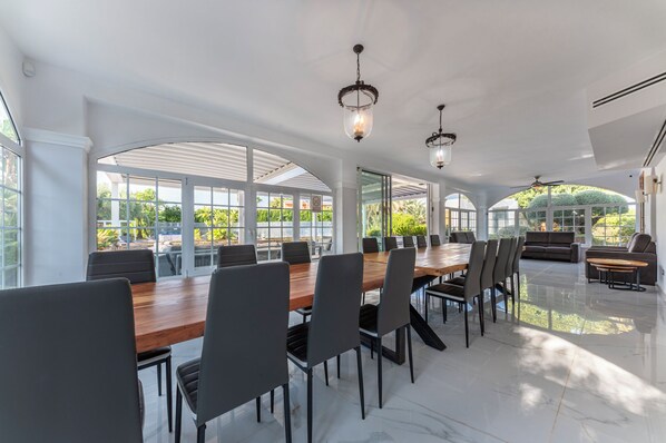 Un vaste espace dédié à la salle à manger, avec une longue table en bois et des chaises modernes, de grandes fenêtres offrant une vue sur la verdure, et des luminaires suspendus élégants.