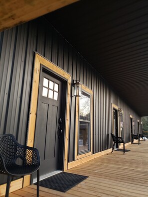 A covered wooden deck with a black door and windows, a black chair, and a wooden bench. - Hidden Jewell Studio Suite #1 - 2BD/1BA Kitchenette & Washer/Dryer on Outlaws  (Lenore)