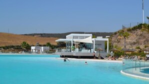 Zona de piscina con un edificio moderno identificado como "LA CAYENA", rodeada de sillas de descanso y sombrillas.