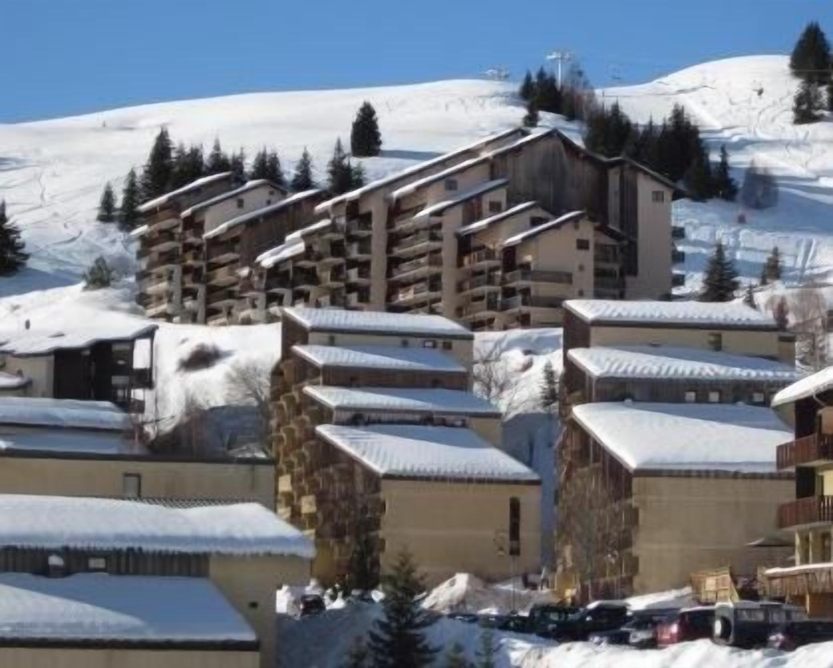 Un complejo turístico en la montaña cubierto de nieve, con varios edificios, incluyendo un hotel grande con balcones y un telesilla al fondo.