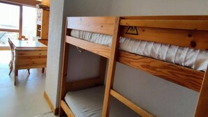 Un lit superposé avec une structure en bois et un matelas, une table en bois avec des chaises, et une fenêtre donnant sur l’extérieur.