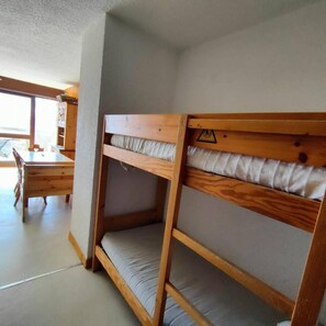 A bunk bed with a wooden frame and a mattress, a wooden table with chairs, and a window with a view outside.