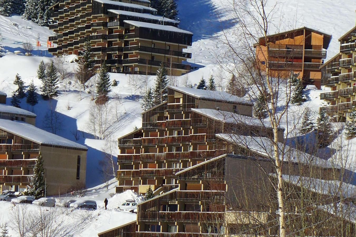 Une station de ski avec des bâtiments à plusieurs étages, des toits enneigés et un paysage montagneux en toile de fond.