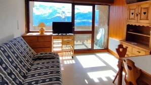 Un salon avec un canapé, des meubles en bois, un téléviseur et une vue sur les montagnes, visible à travers de grandes fenêtres.