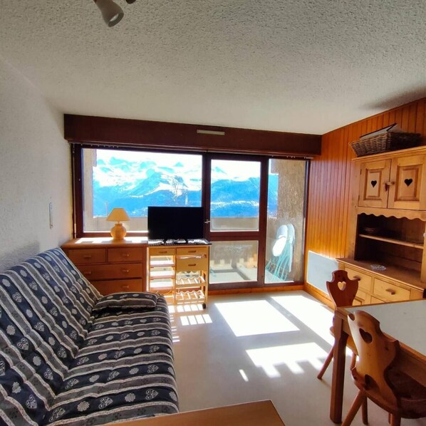 Ein Wohnzimmer mit einer Couch, Holzkästen, einem Fernseher und Blick auf Berge durch große Fenster.