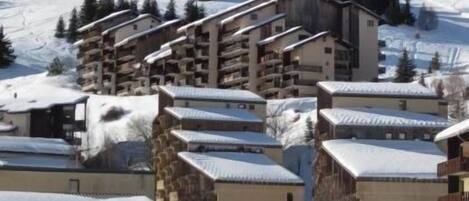 A snow-covered mountain resort with multiple buildings, including a large hotel with balconies and a ski lift in the background.