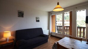 A cozy living room with a dark blue sofa, a wooden dining table, and a hanging lamp. There are two windows with a view of a balcony and greenery outside. - 50m from resort center, Ground floor, view mountain, balcony, parking, ski locker, 23m² (Auris)