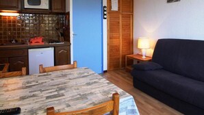 A compact living space with a dining table, chairs, a sofa, a lamp, and a kitchen area with a microwave and a clock. - 50m from resort center, Ground floor, view mountain, balcony, parking, ski locker, 23m² (Auris)