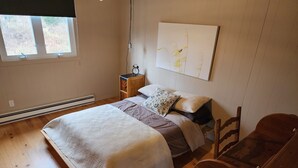 A bedroom with a bed, wooden floor, a nightstand with a clock, a chair, and a window with blinds.
