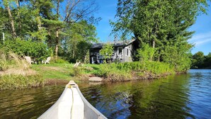 A canoe on a calm river with a house and trees in the background.