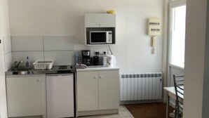 Une cuisine compacte avec des meubles blancs, un four à micro-ondes et une machine à café. Une fenêtre laisse entrer la lumière naturelle et un radiateur est fixé au mur.