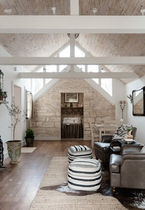 Un loft spacieux avec de hauts plafonds, des poutres apparentes, une cheminée en pierre et un coin salon confortable agrémenté de poufs et d’un canapé en cuir.