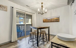 Zona pranzo con tavolo e sgabelli, un lampadario, un quadro e vista attraverso una porta in vetro.