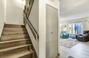 Una scala che conduce a una stanza con una porta, una zona living con un divano e una poltrona, e una finestra con tende.