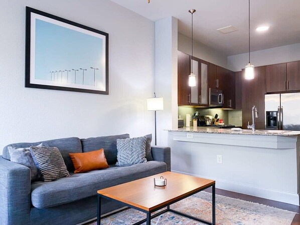A modern living room with a grey sofa, a wooden coffee table, and a kitchen area with dark cabinets and a white countertop.