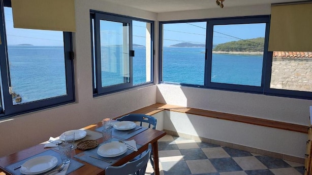 A dining area with a table set for two, large windows with a view of the sea, and a checkered floor. - Apartments Blaža (Prvic Luka)