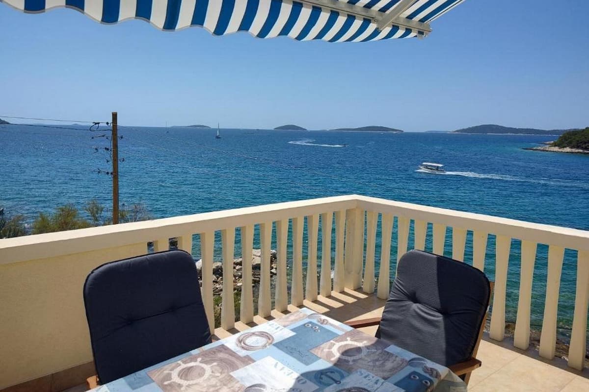 A balcony with two chairs and a table, overlooking a clear blue sea with boats and distant islands.