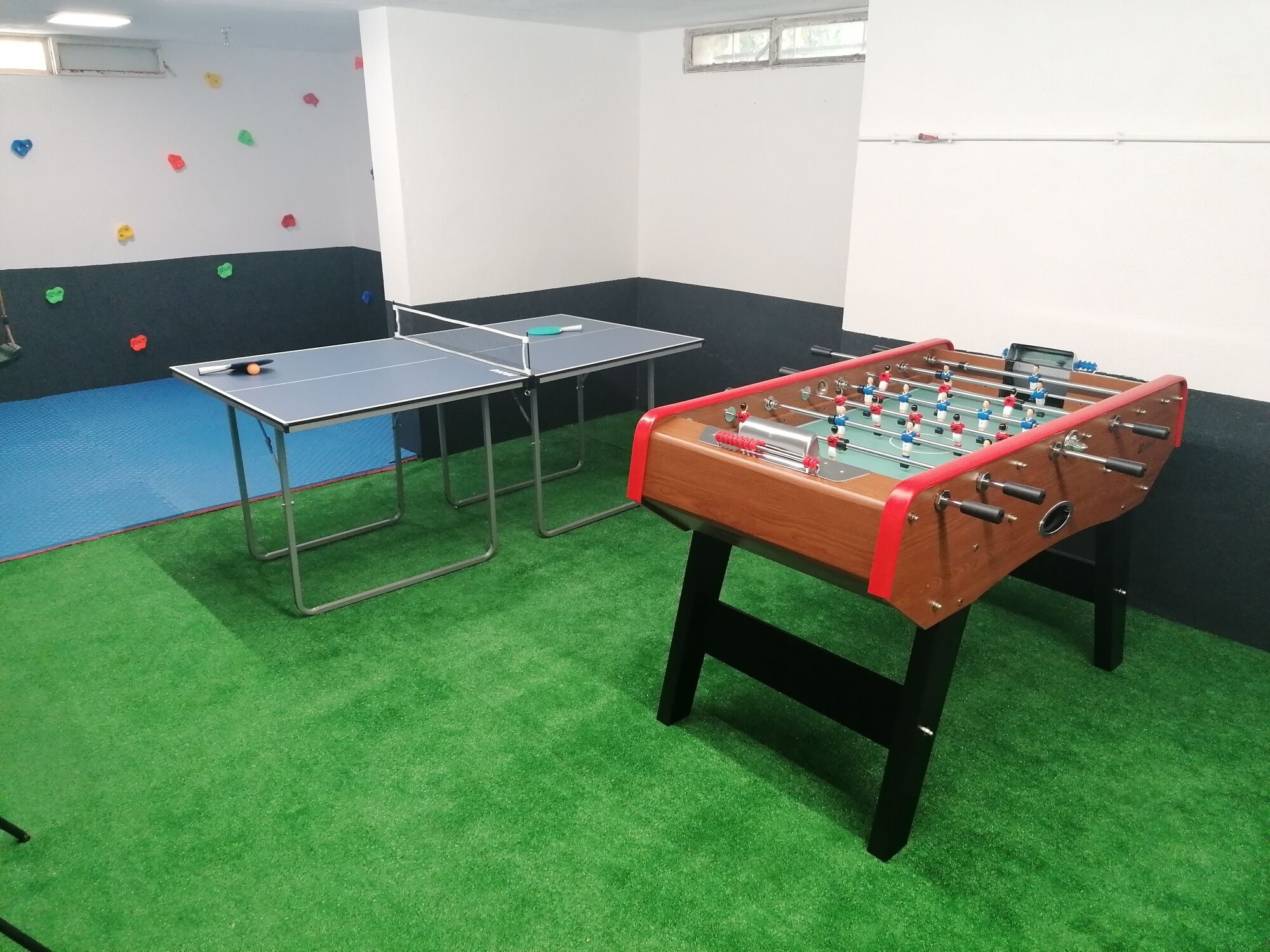 A room with a foosball table, ping pong table, and a foosball table with a red frame.