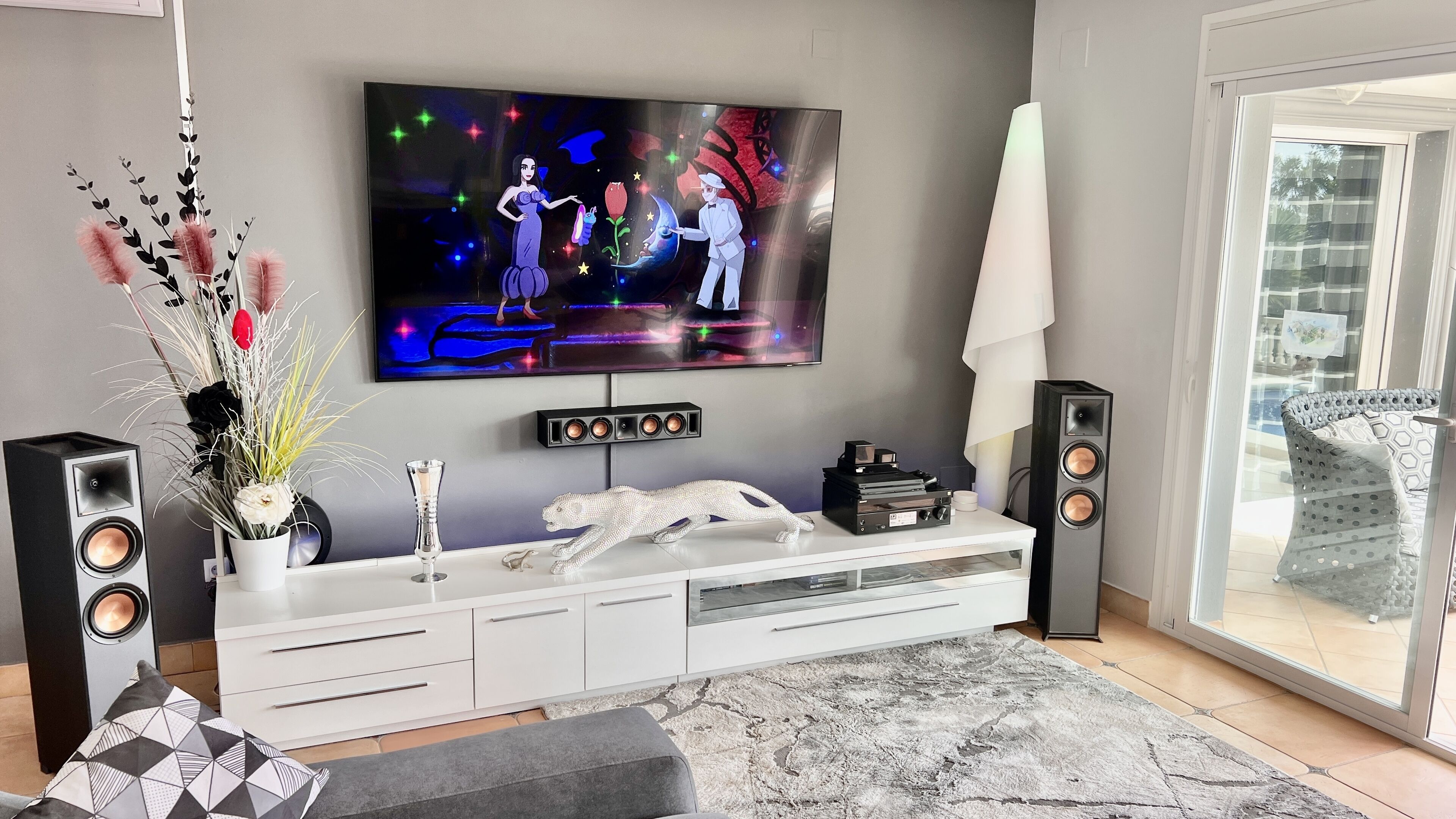 A modern living room with a flat-screen TV mounted on the wall, a white entertainment unit with speakers, a decorative plant, and a patterned rug.