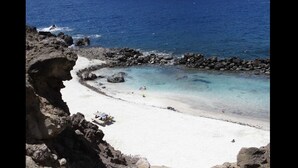 Una playa con agua turquesa cristalina, costa rocosa y algunas personas disfrutando la arena.