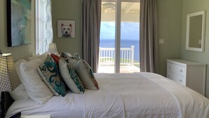Un dormitorio con una cama grande, una mesita de noche, una lámpara, un cuadro con una imagen de un perro y vistas al océano a través de una puerta corrediza.