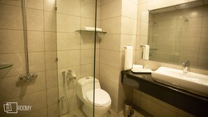 A bathroom with a glass-enclosed shower, a toilet, a sink with a mirror, and a shelf above the toilet. - Roomy Signature Hotel  (Islamabad)