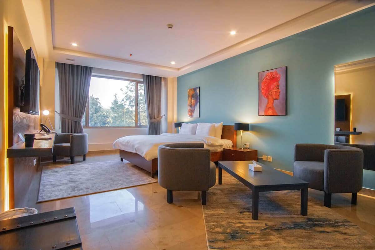 A modern hotel room with a large bed, two armchairs, a coffee table, and a desk with a lamp.
