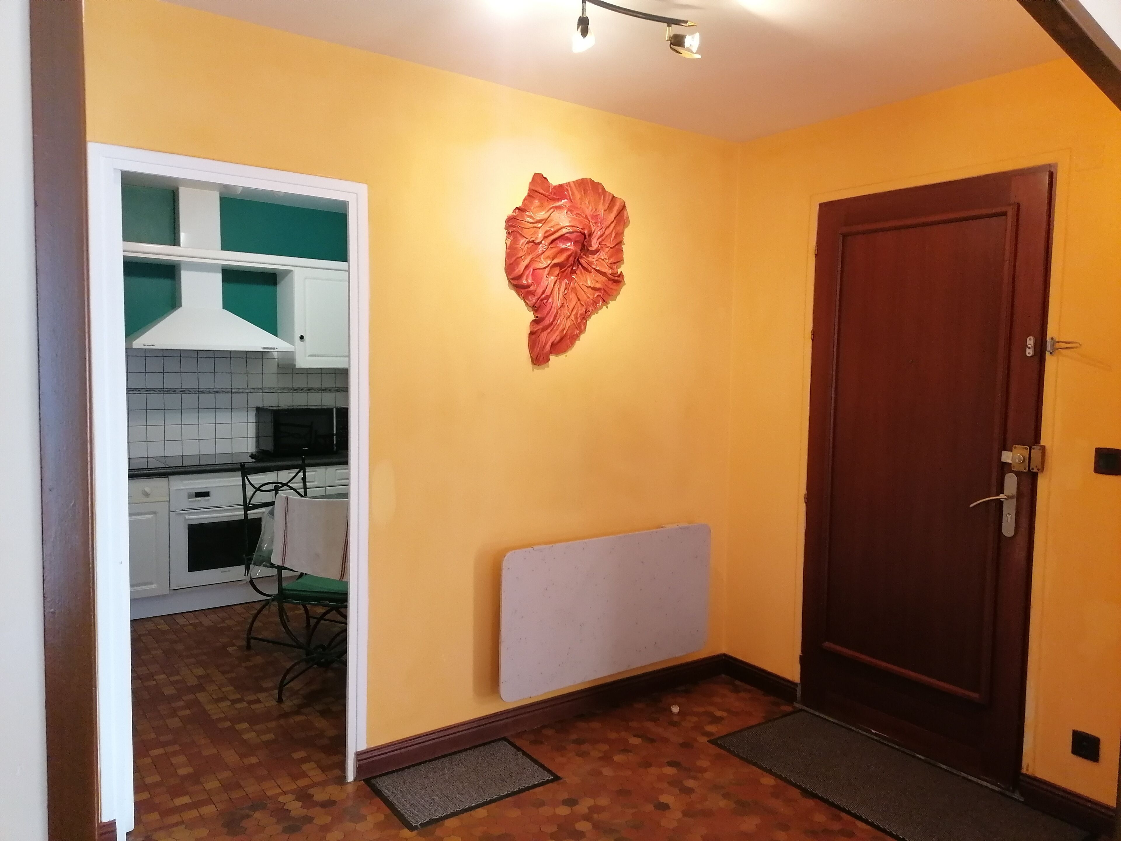 A room with a wooden door, a radiator, a wall-mounted decorative item, and a glimpse of a kitchen area with white cabinets and a green chair.