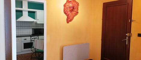 A room with a wooden door, a radiator, a wall-mounted decorative item, and a glimpse of a kitchen area with white cabinets and a green chair.