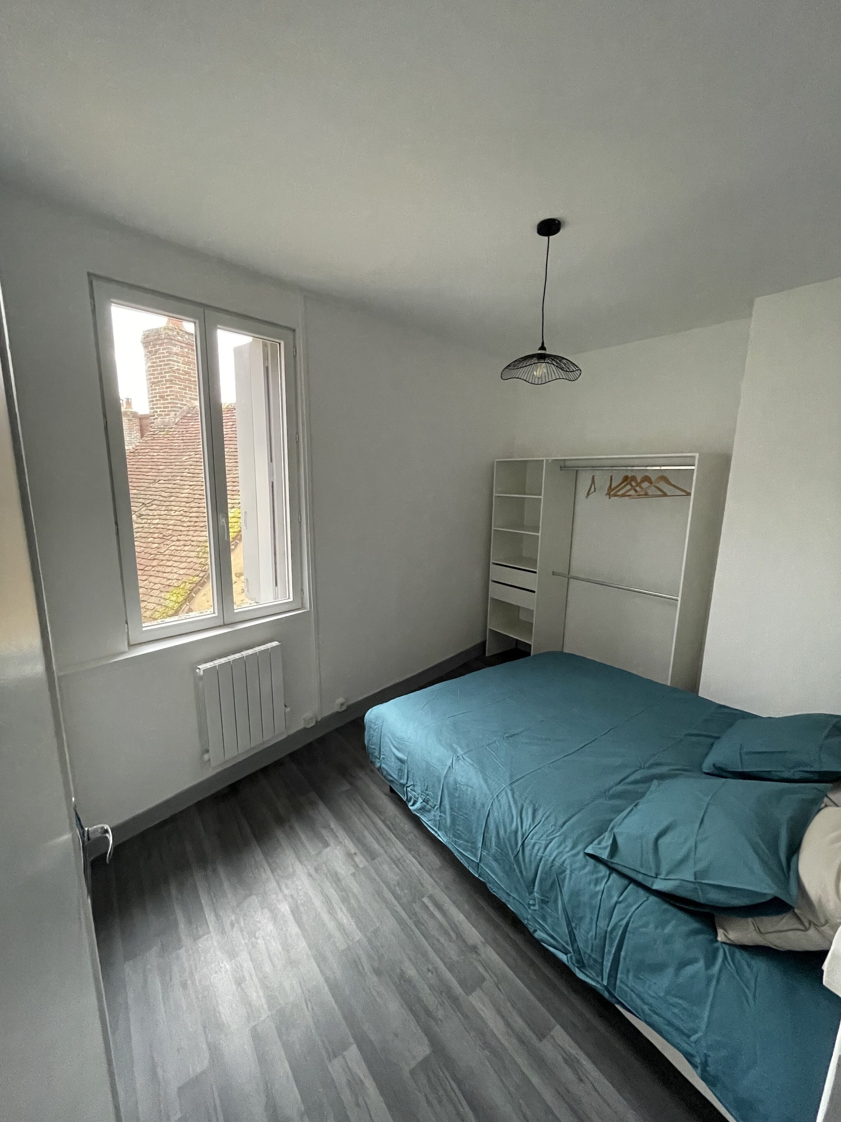 Un dormitorio con un ventanal grande, una cama con ropa de cama azul, un armario blanco y una lámpara colgante.