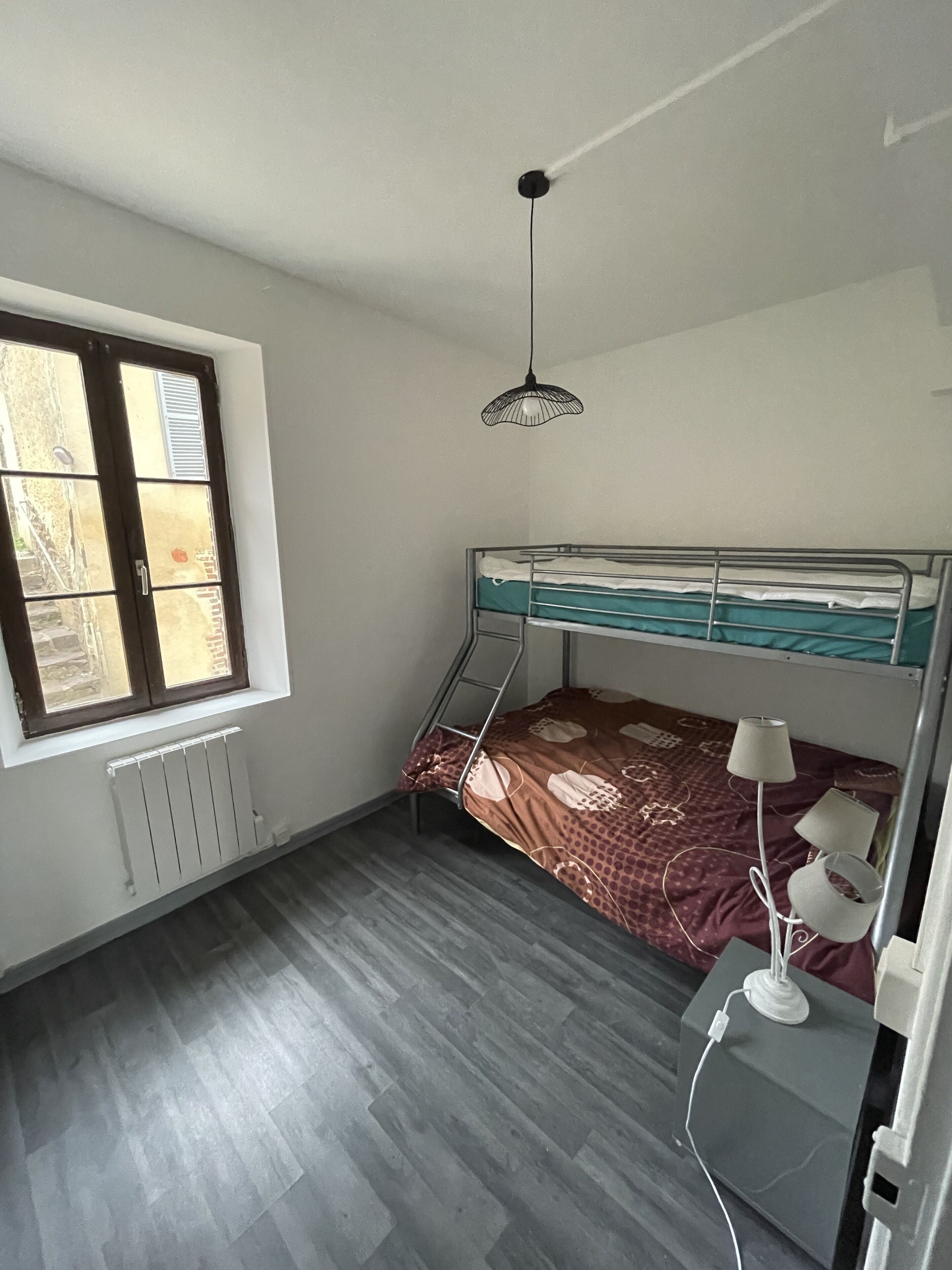 Une chambre avec un lit superposé, un radiateur, une fenêtre avec des stores et un luminaire suspendu.