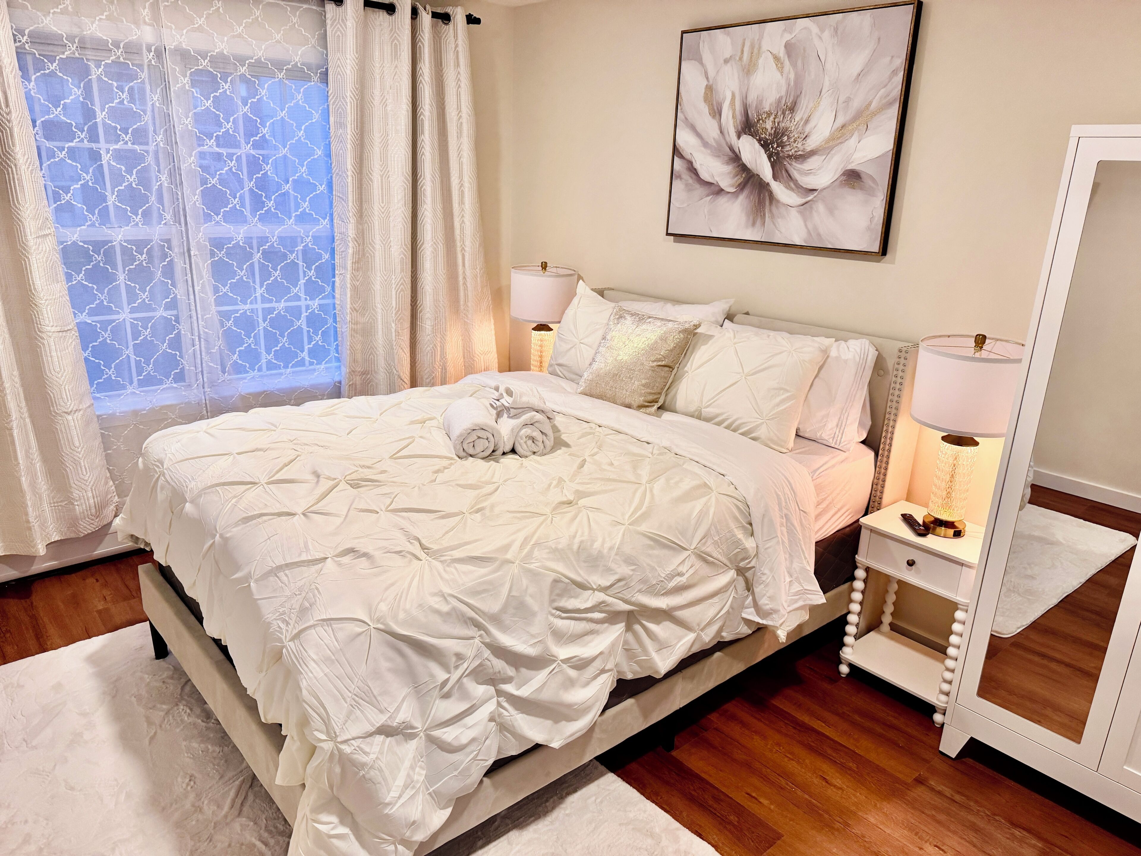 Un letto rifatto con un comofort bianco e cuscini, un comodino con una lampada, e un'ampia finestra con tende a motivi.
